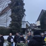 Eine Gruppe von Menschen versammelt sich im Freien, um an einer Veranstaltung teilzunehmen, umgeben von Bäumen und Gebäuden.