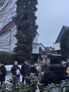 Eine Gruppe von Menschen versammelt sich im Freien, um an einer Veranstaltung teilzunehmen, umgeben von Bäumen und Gebäuden.