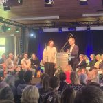 Bei einer Veranstaltung in Hemer spricht ein Mann am Podium, während eine Frau neben ihm steht und das Publikum im Saal aufmerksam zuhört.